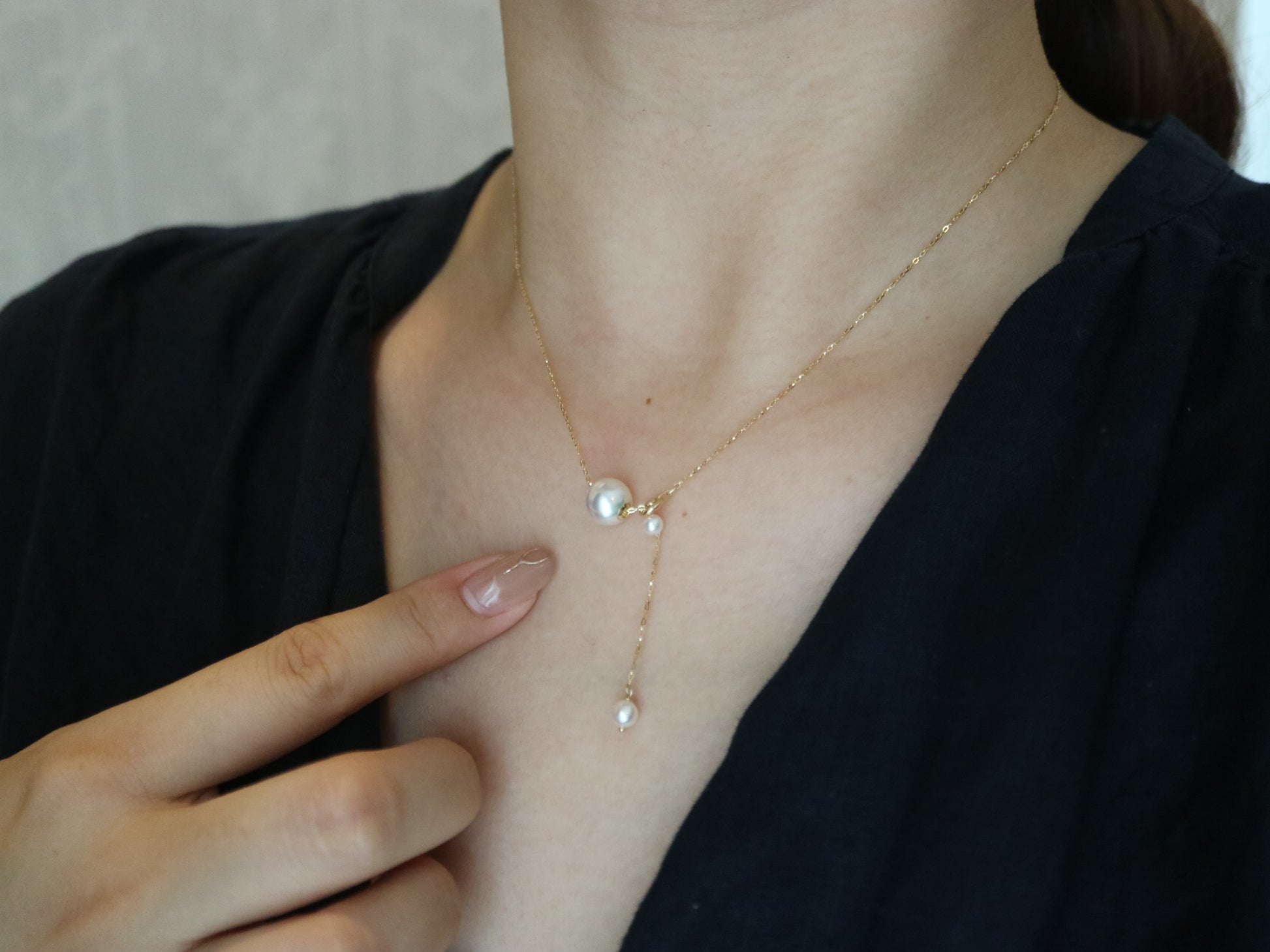 Close-up of a woman wearing a gold chain necklace with three pearls, pointing at it, in black top
