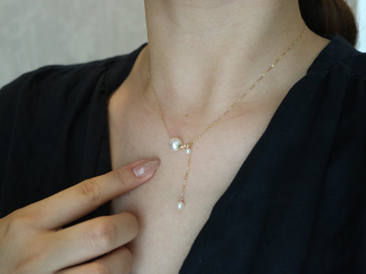 Close-up of a woman wearing a gold chain necklace with three pearls, pointing at it, in black top