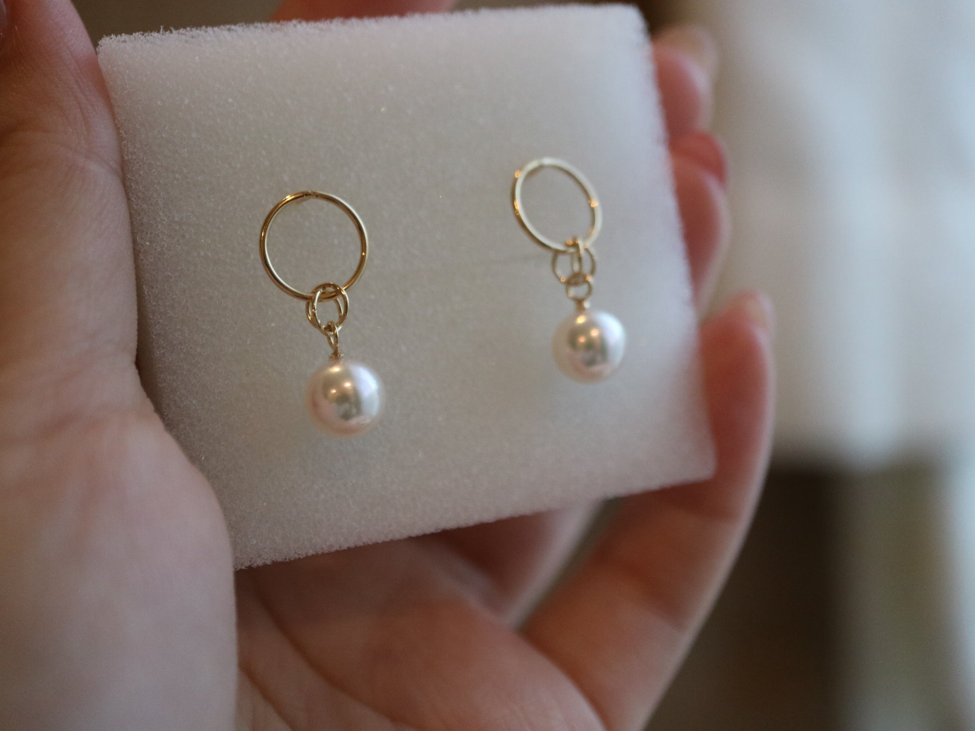 Hand holding gold hoop earrings with white Akoya pearls on a white foam display.