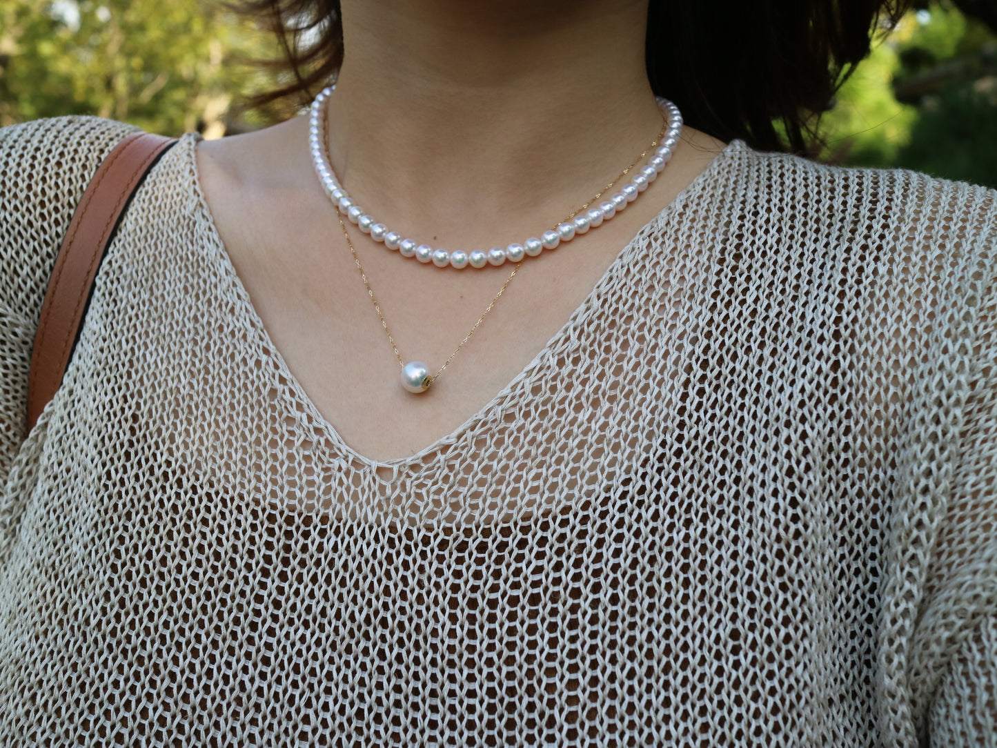 Close-up of woman wearing pearl choker and single pearl pendant necklace with beige mesh top outdoors