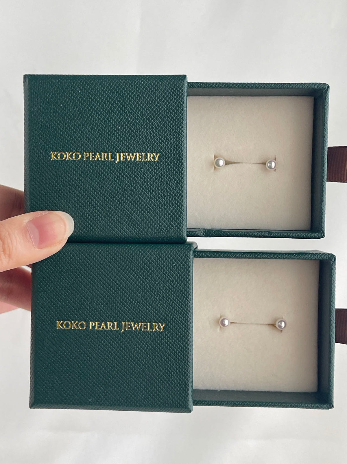 Handheld KOKO PEARL JEWELRY boxes displaying elegant Akoya pearl stud earrings in green textured cases